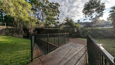 Photo shows a small footbridge that spans the Tarban Creek, situated just to the left of Batemans Road. The left hand of the frame shows lush green grass, as well as some branches hanging over from a tree that is out of frame. The right hand side shows the other side the footbridge, as well as a tree and an incline that leads up to Batemans Road. The pathway that connects to the footbridge then diverts to the left, leading further into the reserve. The sky has wonderful sun with a few storm clouds gathering.