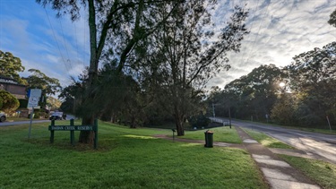Photo shows the eastern accessway into Tarban Creek Reserve. There is a sign situated to the left of frame denoting the location. To the right of frame, you can see Manning Road and the footpath that runs alongside it. Middle of the frame in the foreground and background is very lush grass, as well as a number of large trees, two of which are in the middle ground and several in the background.